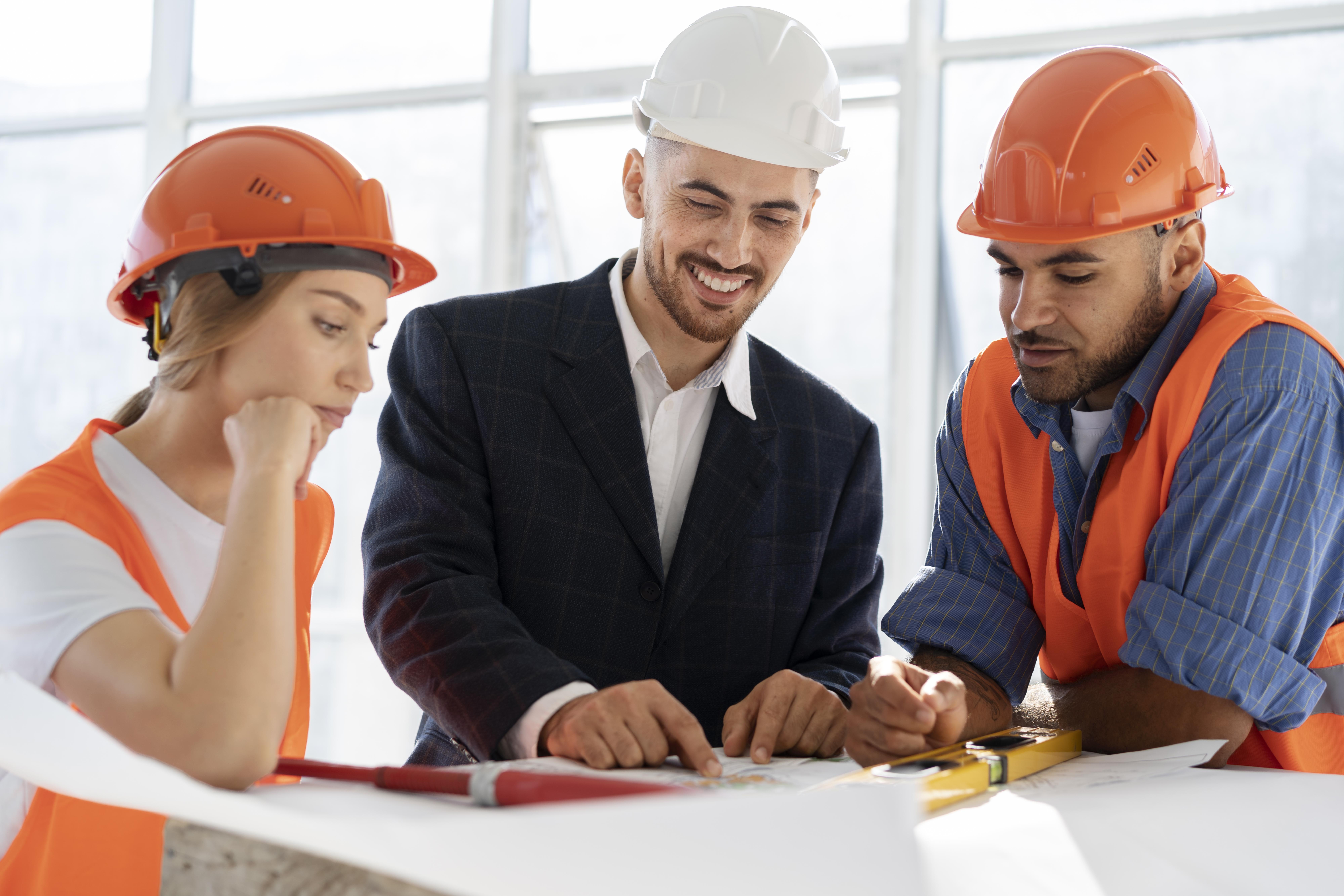 Construction site meeting with two workers in orange safety vests and helmets and a project manager in a suit and white hard hat reviewing plans.