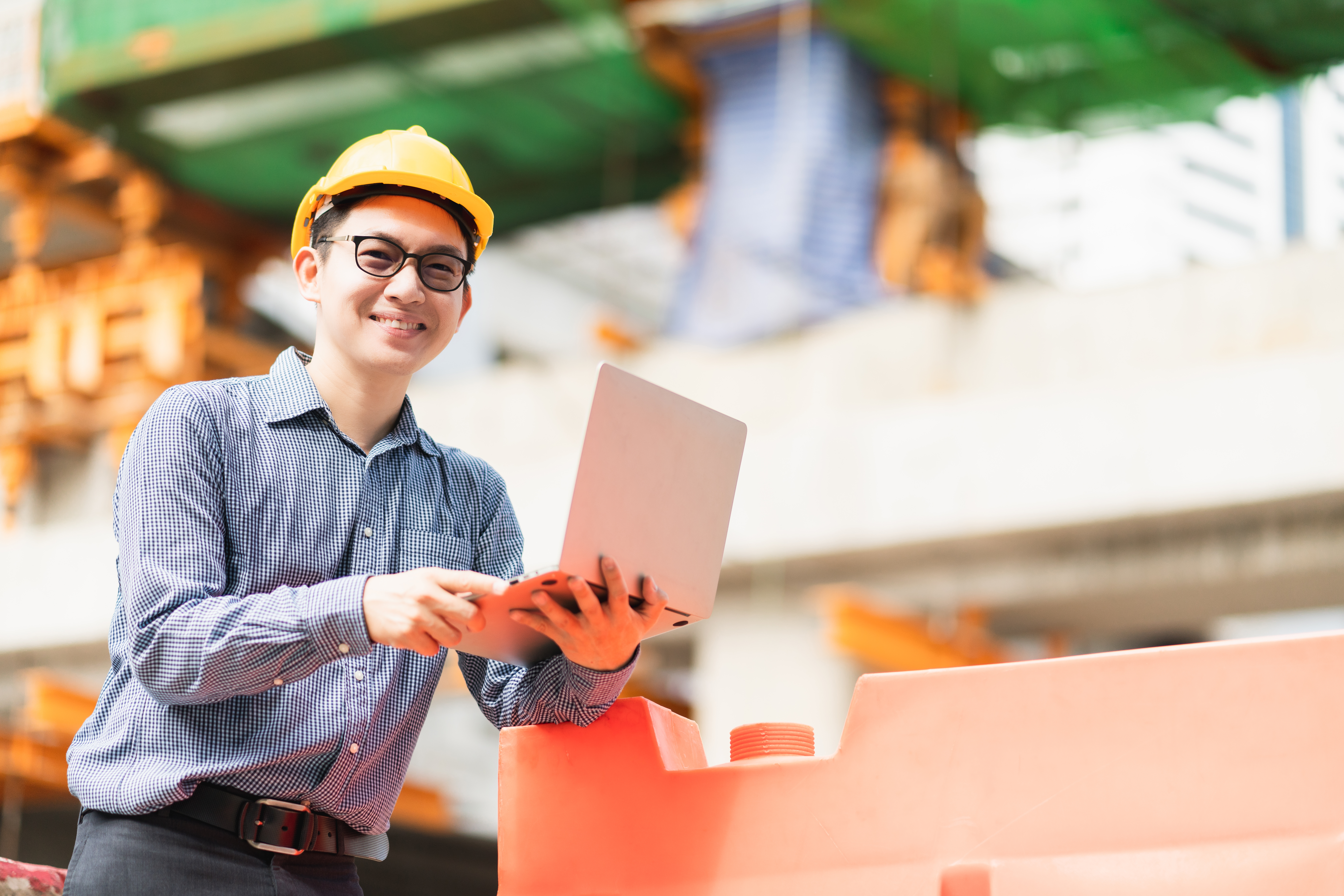 Smiling construction engineer in safety helmet using laptop on active job site to monitor project progress